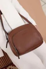 Bandolera de cuero sintético color beige, con diseño compacto y elegante. Cuenta con un bolsillo frontal con cierre y un compartimento principal con cierre. La correa es regulable.