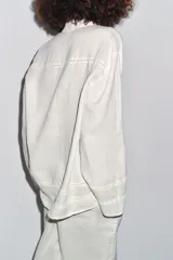 Camisa de lino color crudo, con cuello solapa y escote en pico. Tiene mangas por debajo del codo y detalle de entredós combinados a tono. El bajo es asimétrico y el cierre frontal es con botones.