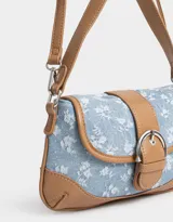 Bandolera de denim celeste con estampado floral blanco, detalles en cuero sintético marrón y correa de hombro ajustable. Cierre con hebilla metálica.