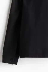 Campera camisera negra de corte recto, con cuello clásico, cierre frontal con cremallera, dos bolsillos de parche con solapa en el pecho y bolsillos laterales.