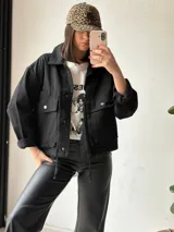 Campera negra de corte recto, con cuello camisero, cierre frontal con botones a presión, bolsillos de parche con solapa en el pecho y mangas largas con puños abotonados.