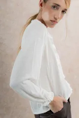 Blusa blanca de lino con cuello con volados, mangas largas con puños elásticos y cierre frontal con botones.