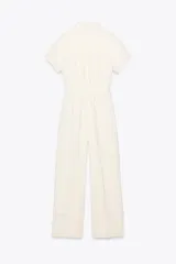 Mono largo color crema, de cuello solapa y manga corta con dobladillo. Tiene bolsillos delanteros y falsos bolsillos traseros. El pantalón es de pierna ancha con pinzas delanteras y dobladillo. Se cierra con cremallera, hebilla y botones.
