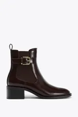 Botín de cuero color marrón oscuro, estilo Chelsea, con elásticos laterales y hebilla decorativa en la caña. Tiene tirador trasero y punta redonda. El tacón es cuadrado y mide 5 cm.