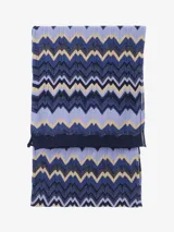 Chal tipo bufanda de punto con patrón de zigzag en tonos azules, lavanda y beige/dorado. Presenta un diseño multicolor con hilos brillantes.