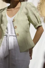 Camisa color verde claro a rayas verticales blancas, confeccionada en lino. Tiene cuello en V, mangas 3/4 abullonadas con puños elásticos y cierre frontal con botones de madera.