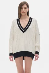 Sweater tejido de punto color crema, con cuello en V con borde negro y doble línea blanca, mangas largas con puños negros, diseño con trenzas en el frente y corte oversize.