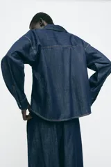 Camisa de denim azul oscuro, corte amplio, cuello solapa, manga larga y cierre frontal con botones a presión.