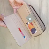 Billetera larga color marrón claro, con cierre de broche a presión en la solapa frontal y cierre de cremallera en el borde lateral. Presenta múltiples compartimentos internos y un bolsillo exterior con cierre.