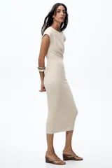 Vestido midi color beige confeccionado en hilatura con algodón. Tiene cuello redondo, manga corta caída y detalle de drapeado lateral.