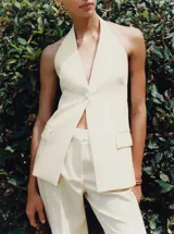 Chaleco largo color crema con cuello halter, espalda abierta, botón joya y bolsillos delanteros.