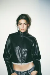 Cazadora corta negra de efecto piel, con cuello subido y manga larga. Tiene bolsillos delanteros con cremallera metálica y cierre frontal con cremallera.