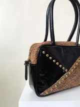 Cartera rígida negra de cuero con tachas plateadas y tres combinaciones de cuero diferentes que aportan textura. Tiene herrajes y cierre metálico plateado marca YKK. Cuenta con un asa fina para llevar al hombro o en la mano y una correa larga extra regulable.