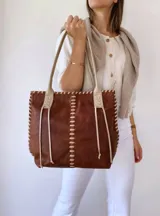 Bolso tipo tote de cuero marrón con costuras blancas a la vista y tiras decorativas. Asas de cuero color crema con detalle de trenzado. Forrada en tela.