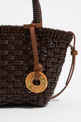Bolso tipo tote de mimbre color marrón oscuro, con doble asa corta y correa de cuero marrón. Incluye dije circular dorado con letras grabadas.