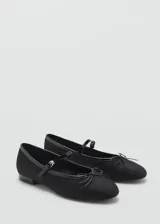 Zapato tipo ballerina color negro con puntera redondeada, tira sobre el empeine con hebilla y moño decorativo.