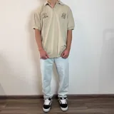 Chomba color beige con cuello clásico, corte oversize, mangas cortas y bordados marrones con la inscripción "Golf Club" y otros motivos. 