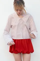 Camisa de popelina a rayas marrones y blancas, con cuello bobo con puntilla y escote pico con lazada. Manga larga con puño y cierre frontal con botones.