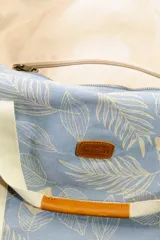 Bolso tipo tote de lona color celeste con estampado de hojas en blanco, detalles en cuero marrón y correa de hombro de lona color crudo.