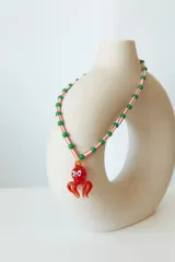 Collar de cuentas de plástico verdes y blancas alternadas, con dije de pulpo rojo de vidrio. El dije de pulpo tiene ojos blancos con pupilas negras.