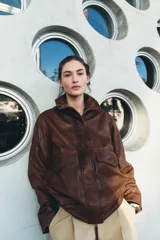 Cazadora bomber de cuero color marrón, con cuello alto, manga larga y bolsillos de parche con solapa en el delantero. Cierre frontal con cremallera metálica y botones a presión ocultos por solapa.