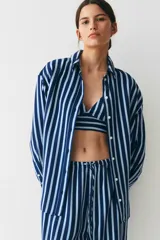 Camisa oversized de sarga a rayas verticales azules y blancas, con cuello de pico y botonadura frontal. Modelo con hombros caídos, mangas largas con tapeta y puños abotonados, y canesú en la espalda. Bajo recto con aberturas laterales redondeadas.