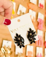 Aretes tipo colgante con diseño asimétrico en forma de hoja o pluma, compuestos por piedras negras facetadas de corte marquesa y pequeñas piedras transparentes brillantes (similares a diamantes o strass). La base es de metal dorado.