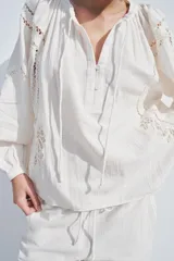 Camisa color crudo confeccionada en mezcla de algodón y lino, con cuello redondo y escote pico ajustable con lazadas. Tiene manga larga abullonada y detalle de bordados perforados a tono.