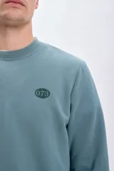 Sudadera verde con cuello redondo y mangas largas. Estampado en la espalda con la frase "Scorpions thrive", un escorpión, el número 073 y un texto descriptivo.