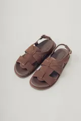 Sandalia plana de gamuza color marrón oscuro, con tiras cruzadas en la parte delantera y una tira fina ajustable con hebilla metálica en el tobillo. Posee plantilla de cuero y suela baja.