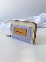 Billetera pequeña con cierre metálico, cuerpo principal de cuero lila texturizado y ribetes en color crema. Presenta un parche de cuero marrón con la palabra "chowie" escrita en cursiva.