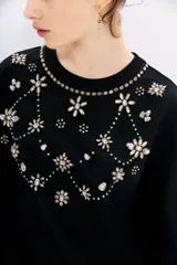 Buzo negro de algodón con cuello redondo y mangas largas. Presenta apliques bordados de piedras brillantes en forma de flores y otros motivos decorativos en el frente.