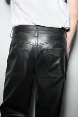 Pantalón de cuero negro, de tiro medio, con trabillas en la cintura, cinco bolsillos, pespuntes marcados a tono, pierna recta y cierre frontal con cremallera y botón.