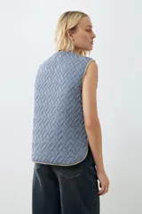 Chaleco capitoneado reversible a rayas azules y blancas con ribete color crema. Tiene cuello redondo, cierre con botones a presión y bolsillos de parche. El reverso es liso color tostado.