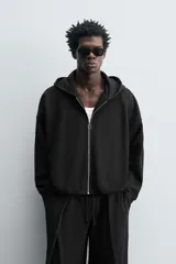 Sudadera negra con capucha, corte boxy, cierre frontal con cremallera, bolsillos laterales y puños elásticos.