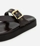 Sandalias planas de PU color marrón oscuro, con tira entre los dedos y tira ancha sobre el empeine con hebilla dorada ajustable.