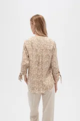 Blusa color beige con estampado de arabescos blancos, cuello mao, mangas 3/4 con lazos en los puños, cierre parcial con botones y bolsillos frontales.