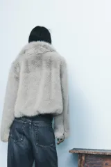Abrigo corto de pelo sintético color gris claro, con cuello subido y manga larga. Tiene bolsillos laterales ocultos en la costura, forro interior combinado y cierre frontal con ganchos metálicos forrados.