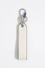 Llavero de cinta de piel color beige claro con logo en relieve y forro interior color azul eléctrico. Incluye mosquetón y anilla metálicos.