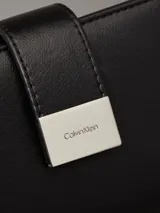 Billetera mediana tipo bifold de piel sintética negra, con cierre de broche a presión y placa metálica con el logo de Calvin Klein en el frente.