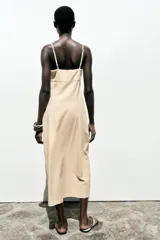 Vestido midi color beige confeccionado en lino, con tirantes finos ajustables, escote en pico con detalle de crochet y jaretas, abertura frontal y cierre lateral con cremallera oculta.