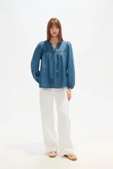 Blusa de lyocell en tono azul denim, con cuello en V, cierre frontal parcial con botones y mangas largas abullonadas con puños elásticos. Presenta un diseño holgado con detalle de fruncido en el pecho.