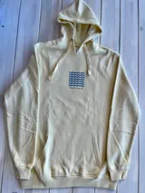 Hoodie de color crema con capucha y puños ajustados. Estampado trasero con un sol estilizado en color terracota y tres olas onduladas en color gris azulado.