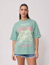 Remera blanca de algodón con estampado frontal de estilo playero con texto "Welcome to the Surf Camp" y dibujo de una casa de playa con palmeras y tablas de surf.
