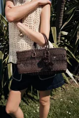 Bolso de mano marrón con cuerpo trenzado y doble asa. Incluye charm extraíble y bolsa interior con cierre fruncido.