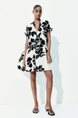 Vestido corto tipo túnica color blanco con estampado de flores negras, cuello mao, mangas cortas con doblez, botones frontales y falda con vuelo. Corte holgado.