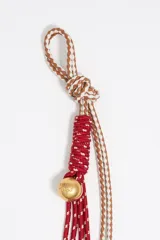 Colgante para bolso con cordones trenzados en color rojo y beige, detalle de cuenta dorada y mosquetón metálico.