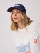 Gorra de lona blanca con visera azul marino y malla trasera azul marino. Presenta un logo bordado en el frente con letras rosadas y contorno azul marino.