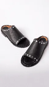 Sandalias de cuero color marrón claro, con punta descubierta y diseño tipo mule. Presentan tachas doradas en el borde superior y suela plana con pequeño tacón. Fabricadas en Uruguay.