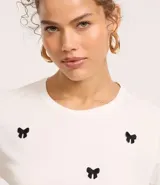 Remera baby look blanca de algodón con cuello redondo y mangas cortas. Presenta bordados de lacitos negros en el frente.
